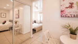A modern hotel room with a glass door leading to a balcony, a bed with a white duvet, a desk with a chair, and a small table with a plant. - El Rancho Motel (Coronado)
