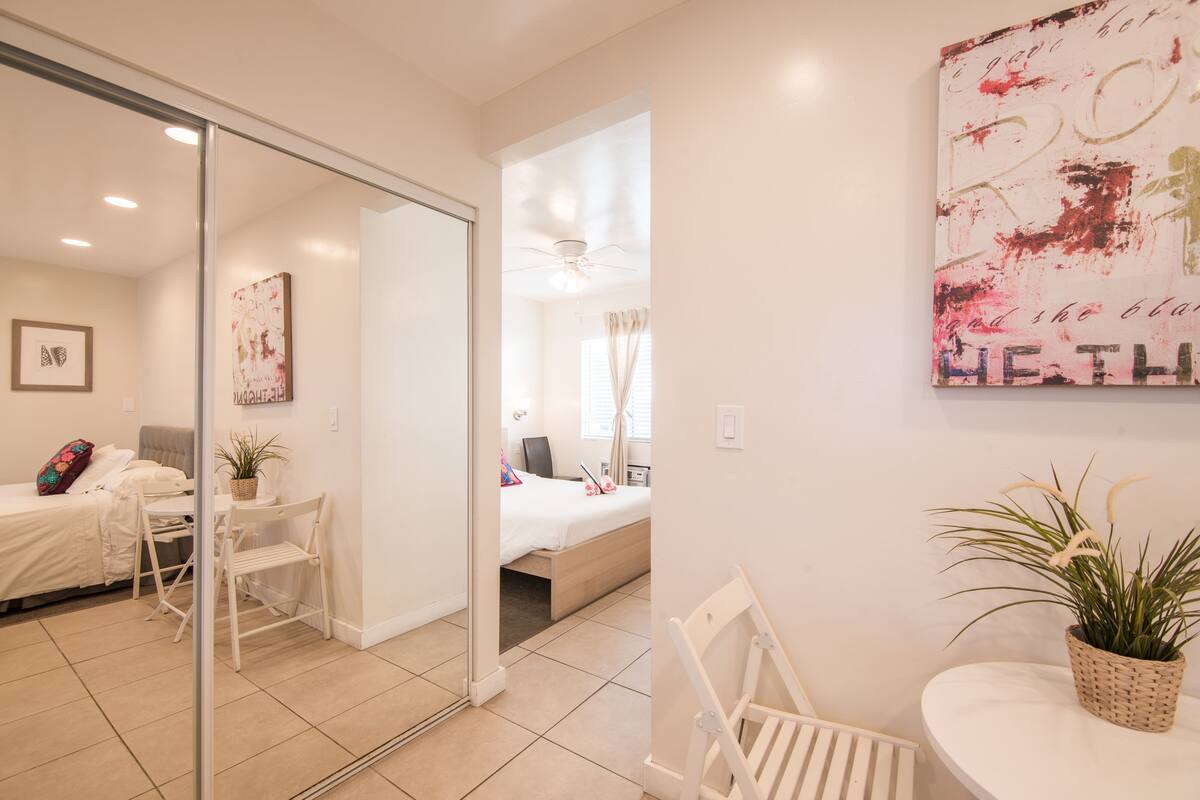 Habitación de hotel moderna con puerta de vidrio que da a un balcón, cama con edredón blanco, escritorio con silla y una mesita con una planta.