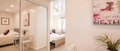 Quarto de hotel moderno com porta de vidro que dá para uma varanda, cama com edredom branco, escrivaninha com cadeira e pequena mesa com planta.