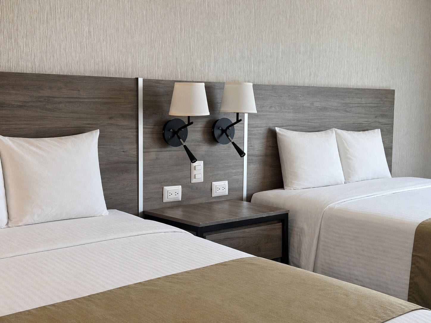 A hotel room with two beds, a nightstand, and two wall-mounted lamps.