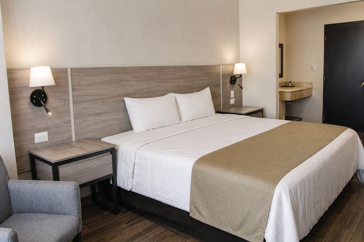 Ein Hotelzimmer mit einem großen Bett, zwei Nachttischen, einem Sessel und einem durch eine offene Tür einsehbaren Badezimmer.