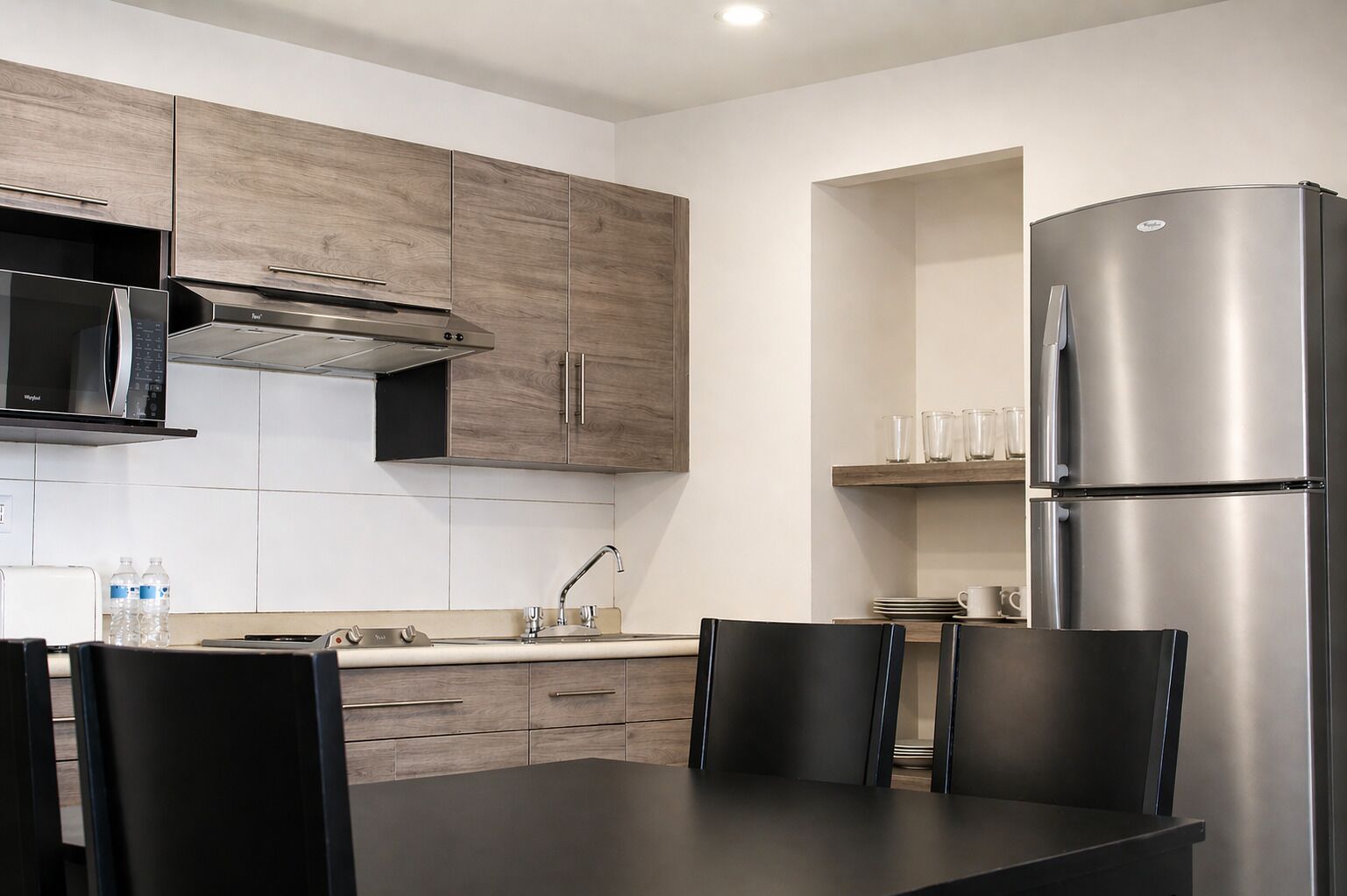 A modern kitchen with wooden cabinets, a stainless steel refrigerator, a microwave, and a dining area with chairs.