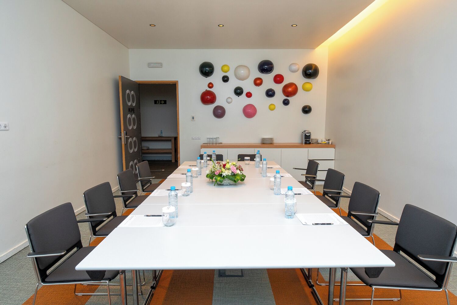 Un salón de conferencias con una larga mesa, sillas, botellas de agua y un centro de mesa floral.