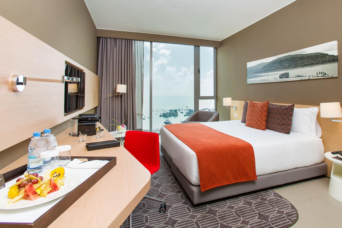 A hotel room with a bed, a desk with a tray of fruit, a TV, and a view of the sea.