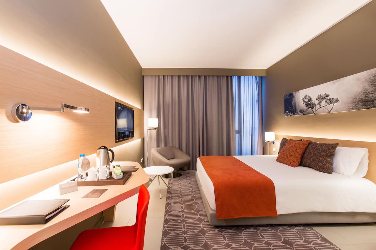 A hotel room with a bed, desk, chair, and a painting on the wall.