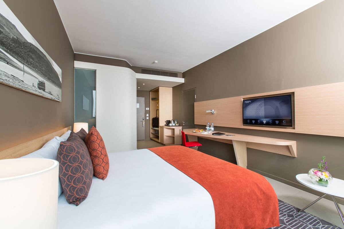A modern hotel room with a bed, a desk, a TV, and a small table with a vase of flowers.