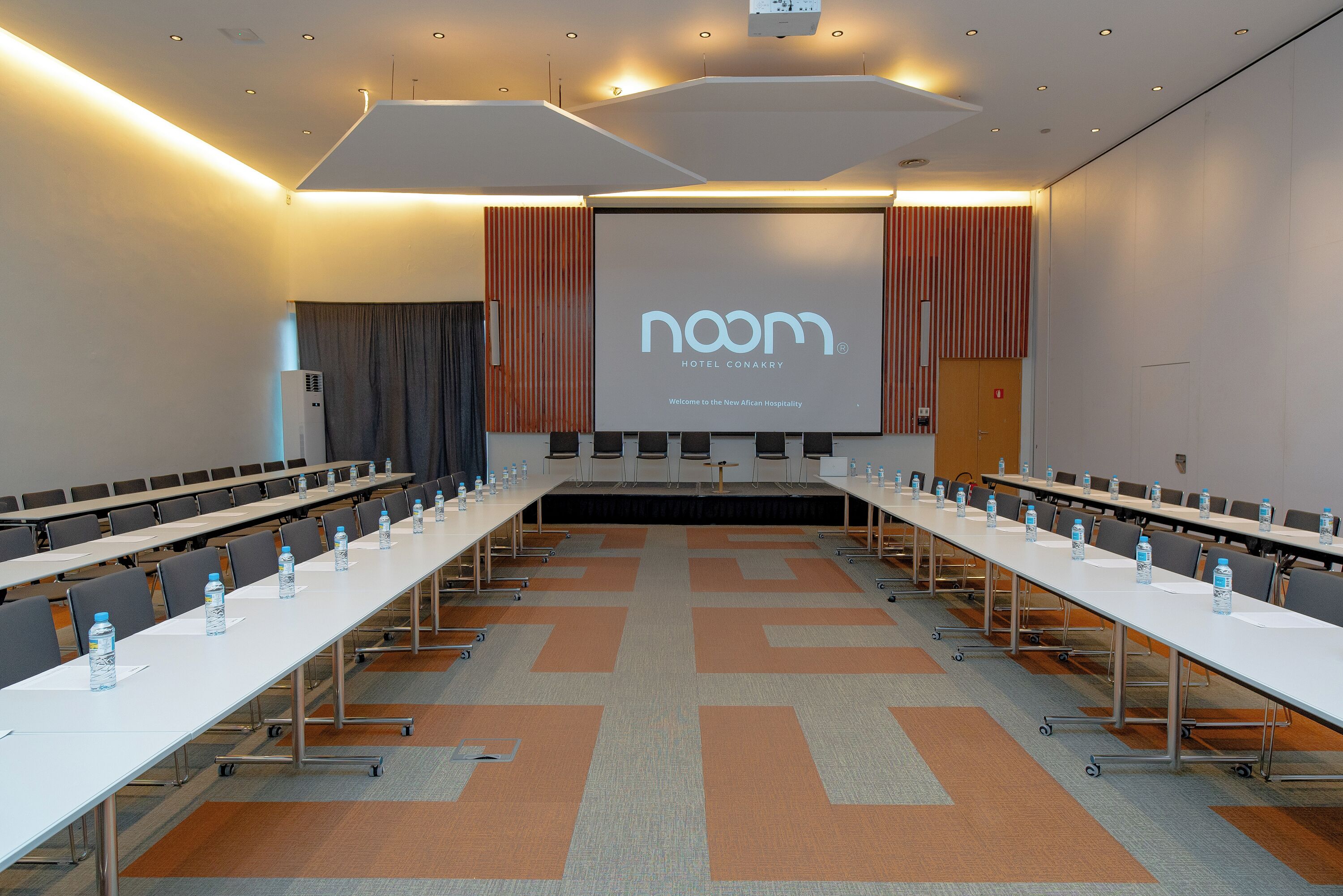 Une salle de conférence avec une longue table, des chaises et un écran affichant le message : « Hôtel Noam, Conakry, bienvenue dans le nouvel univers de l’hôtellerie africaine. »
