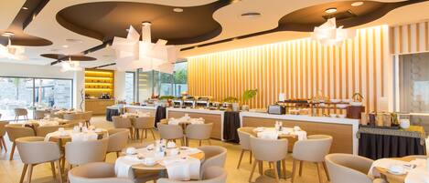 A modern dining area with round tables, white chairs, and a central counter with a variety of food items.