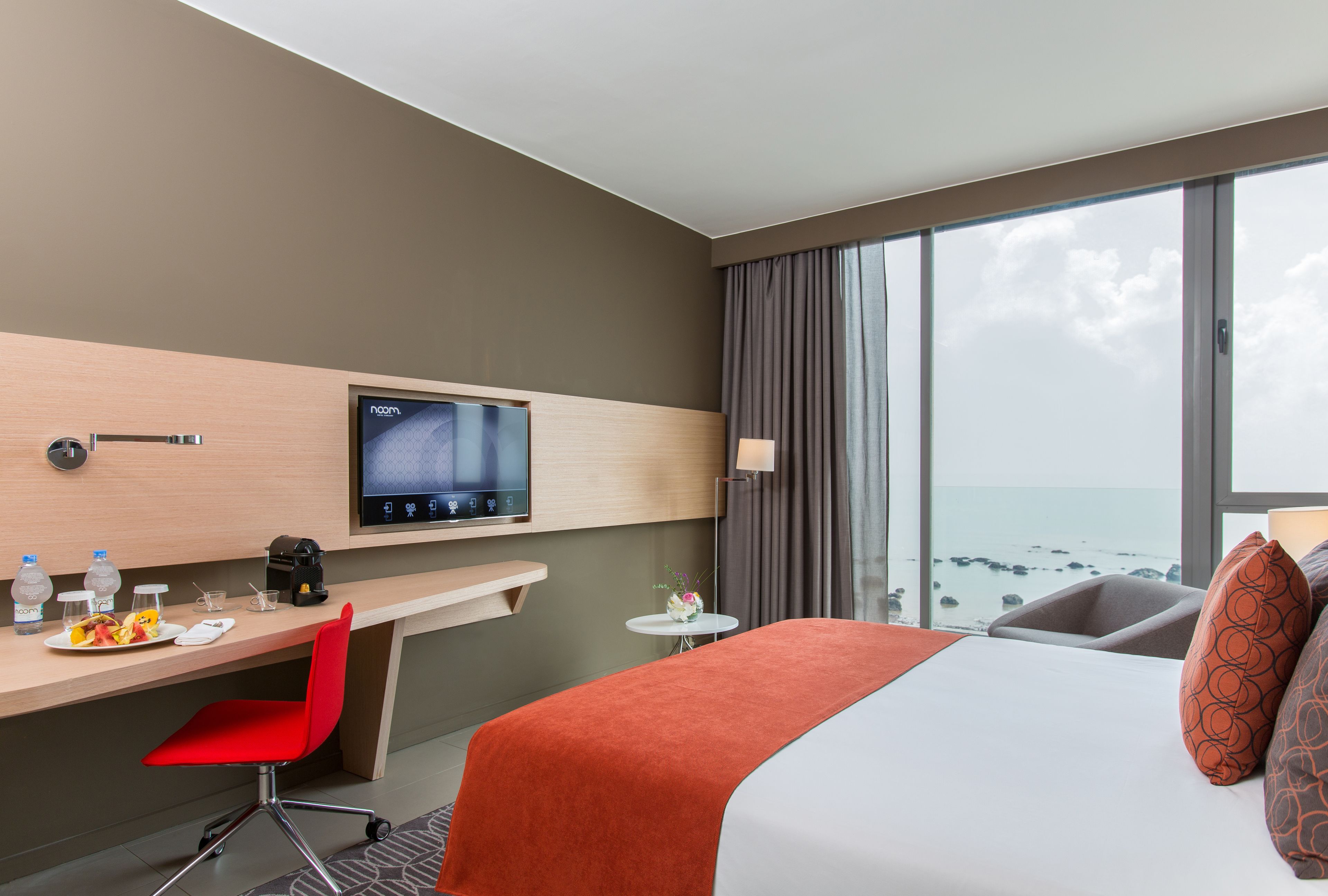 Habitación de hotel con cama, escritorio con silla, televisor, pequeña mesa con lámpara y vista al océano.
