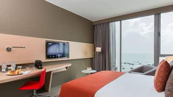 Habitación de hotel con cama, escritorio con silla, televisor, pequeña mesa con lámpara y vista al océano.