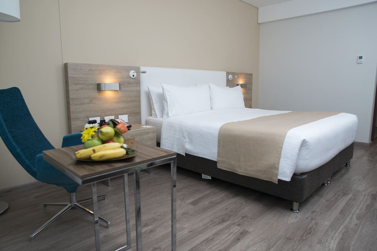 A hotel room with a bed, a desk with a fruit bowl, and a blue chair.
