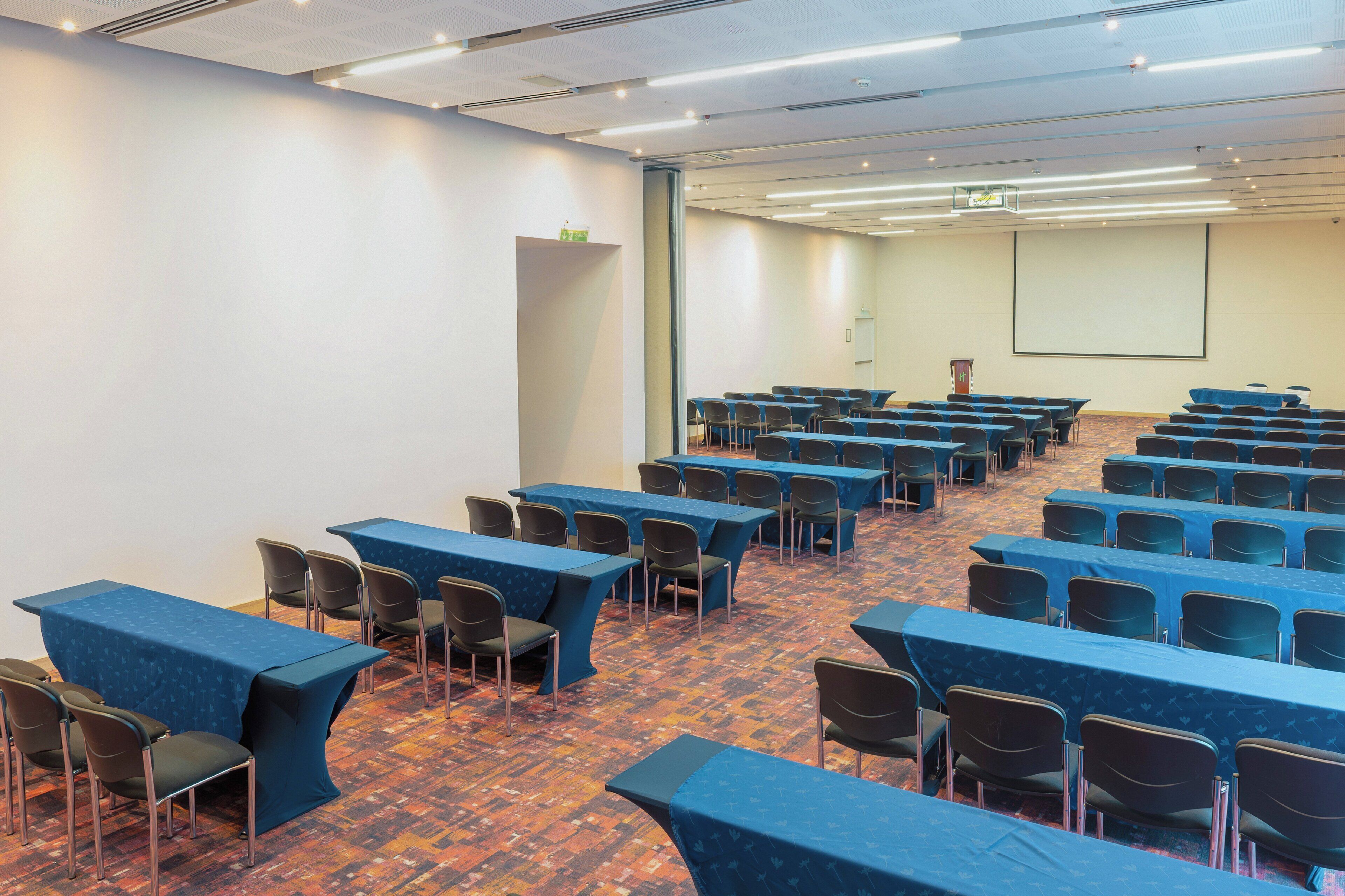 Une salle de conférence avec des rangées de tables et de chaises, un écran de projection et un sol recouvert de moquette.