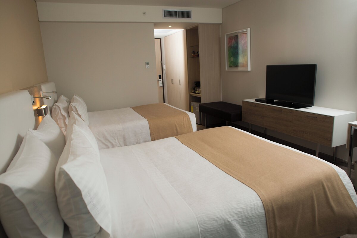 A hotel room with two beds, a desk, a television, and a painting on the wall.