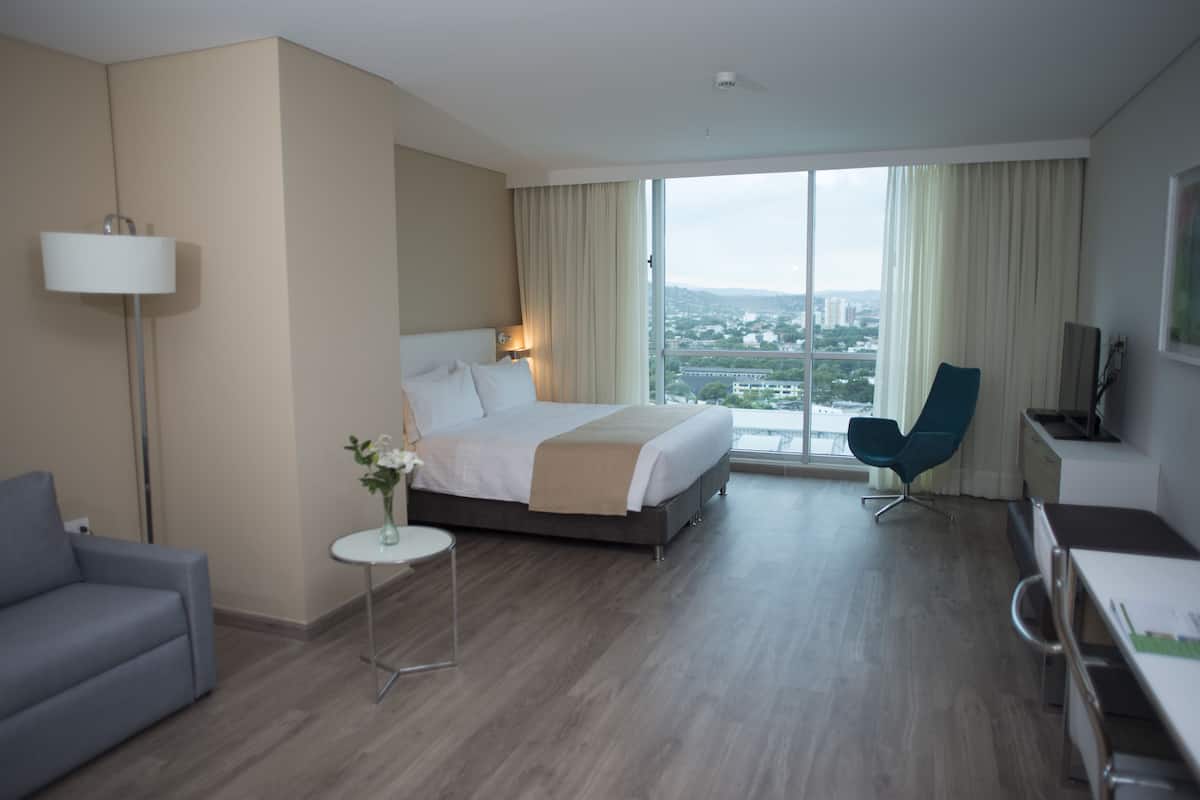 A hotel room with a bed, a desk, a chair, a lamp, and a view of the city.