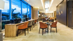 Un moderno vestíbulo de hotel con un mostrador de recepción alargado, varias zonas de estar y grandes ventanales que ofrecen vistas a palmeras y un paisaje urbano.