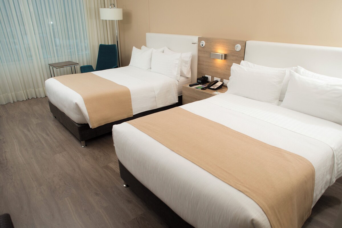 A hotel room with two beds, a bedside table, a lamp, and a curtain.