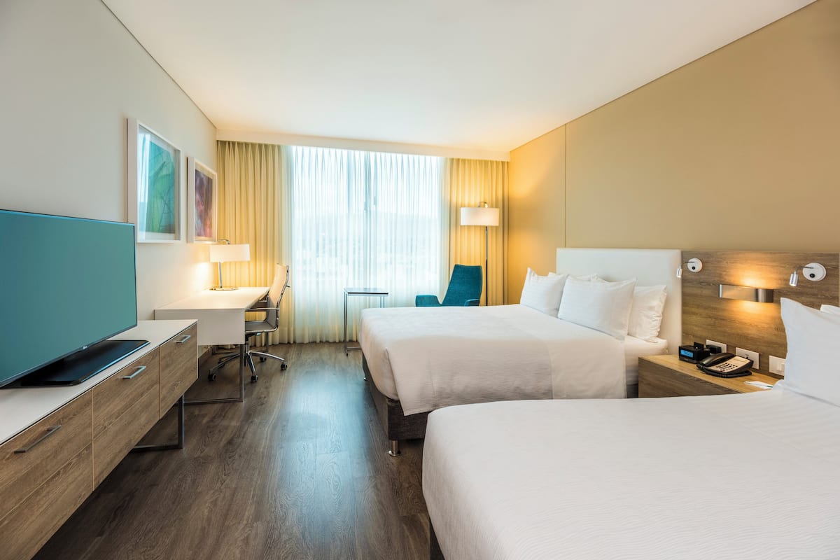 A hotel room with two beds, a desk, a chair, a TV, and a lamp.