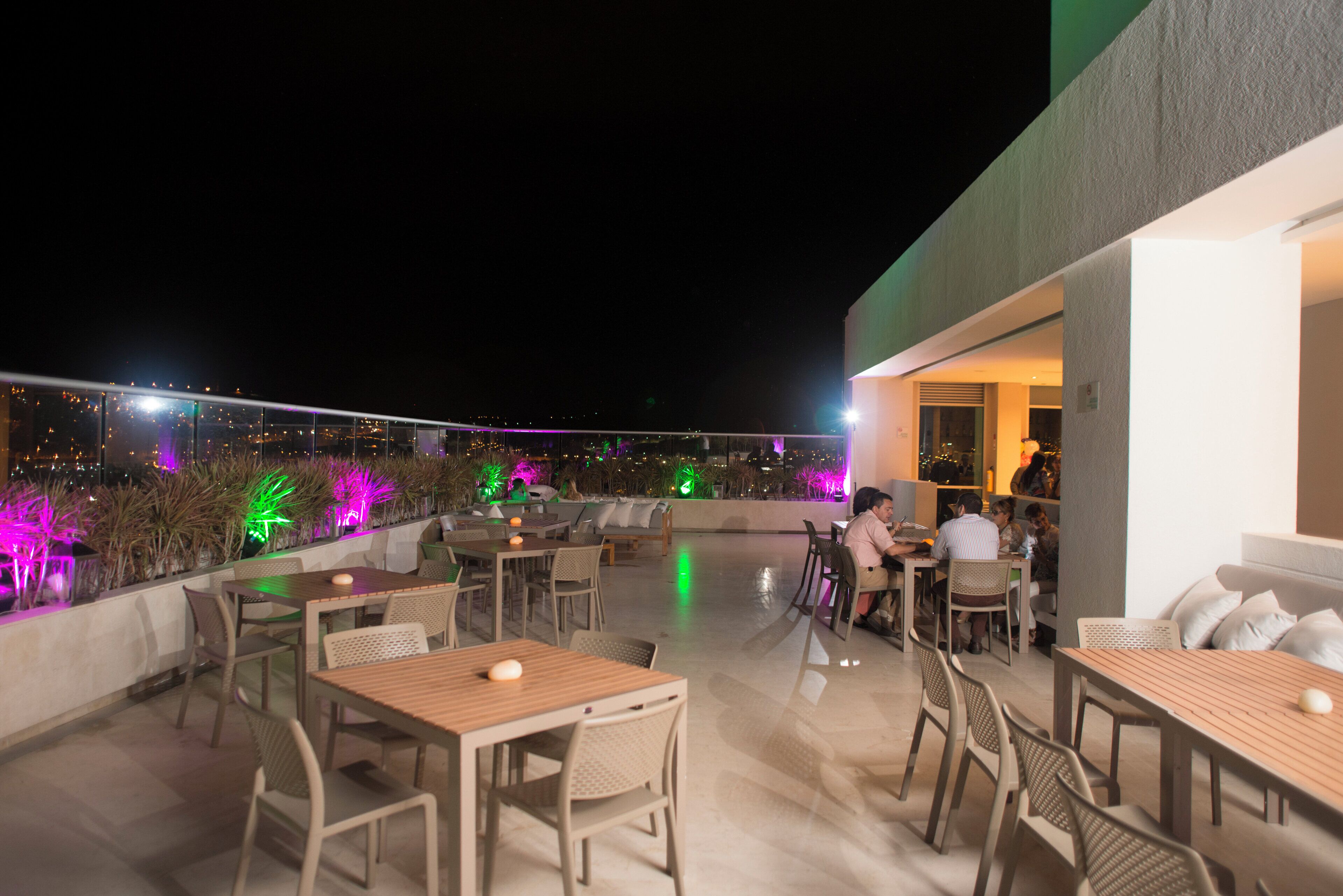 An outdoor dining area with tables and chairs, a view of the city at night, and a building entrance.