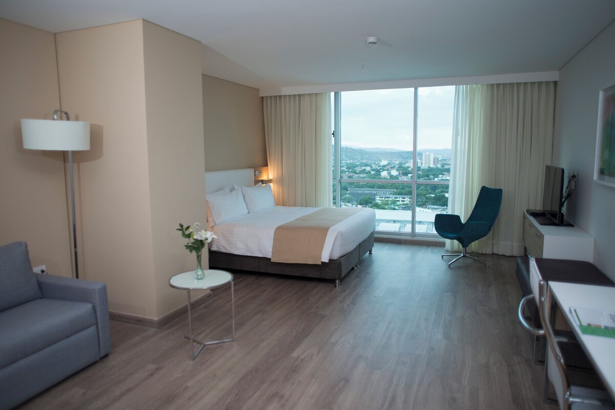 Une chambre d’hôtel équipée d’un lit, d’un bureau, d’une chaise, d’une lampe et offrant une vue sur le paysage urbain grâce à de grandes fenêtres.