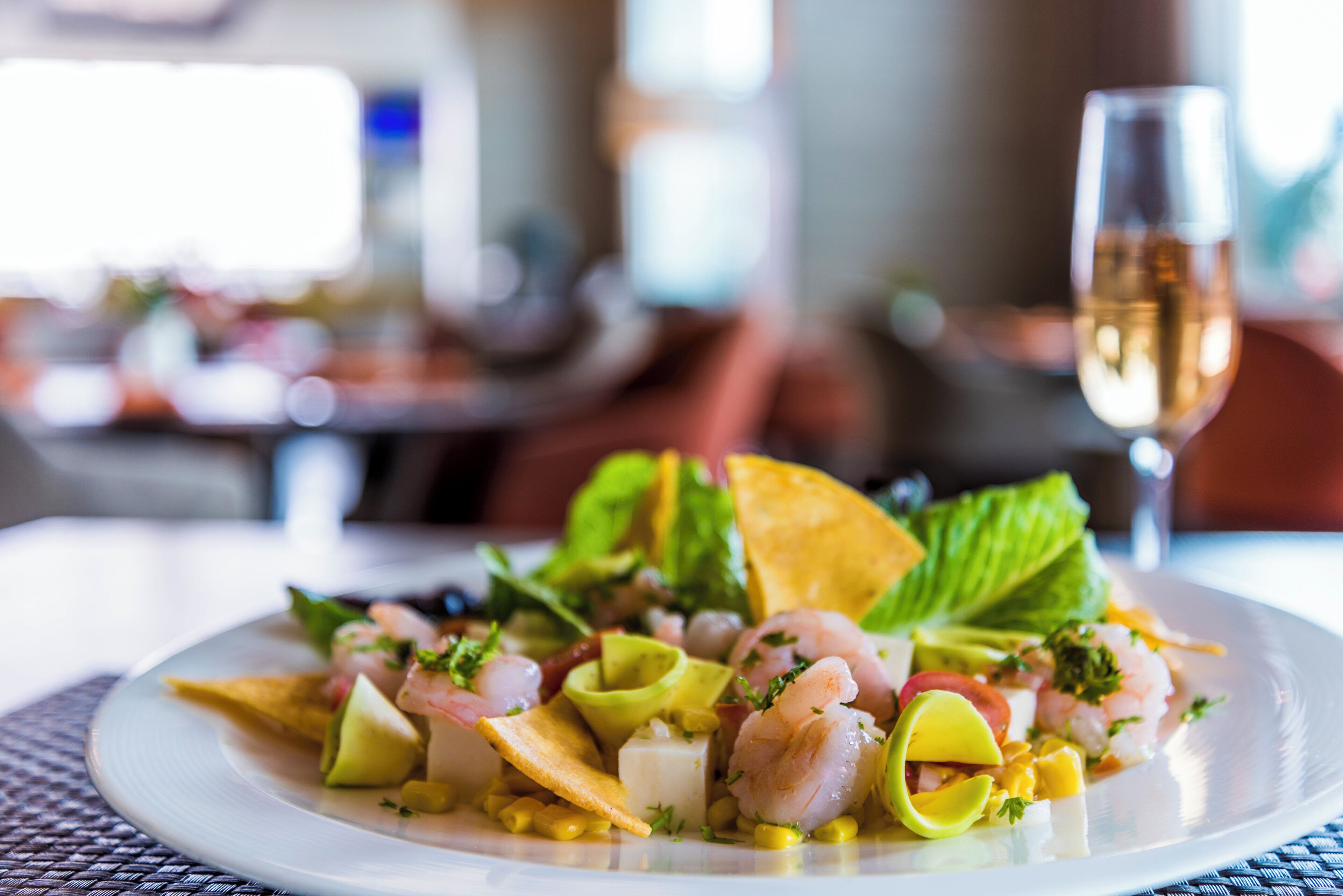 Un plato de ensalada de mariscos con aguacate y maíz, acompañado de una copa de vino blanco, sobre una mesa con un fondo borroso de un restaurante.