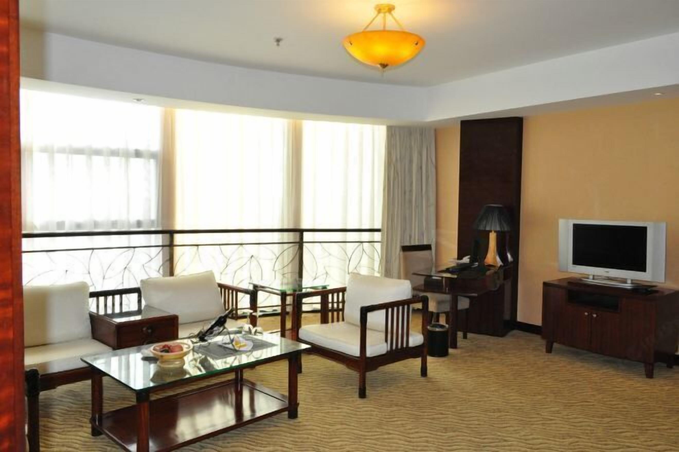 A hotel room with a sofa, armchair, coffee table, TV, and a balcony with a railing.