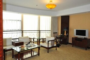 A hotel room with a sofa, armchair, coffee table, TV, and a balcony with a railing.