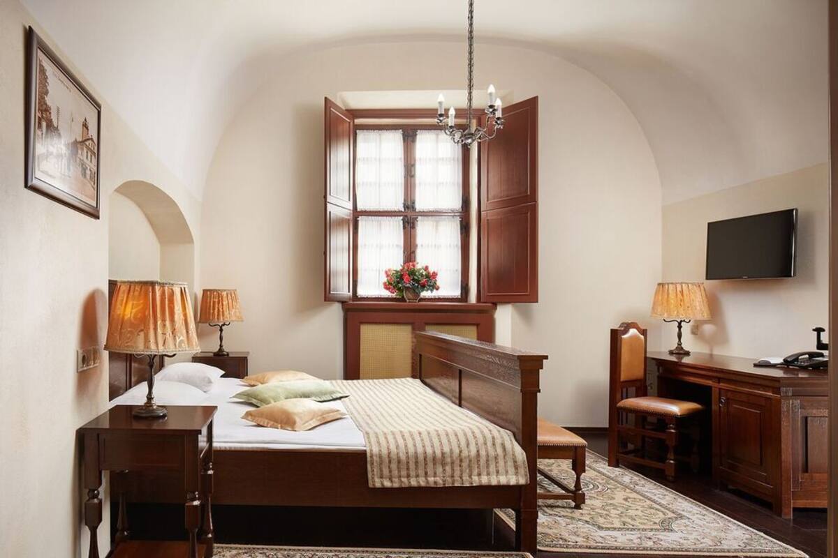 A bedroom with a wooden bed, two bedside tables with lamps, a desk with a chair, a television, and a window with curtains.