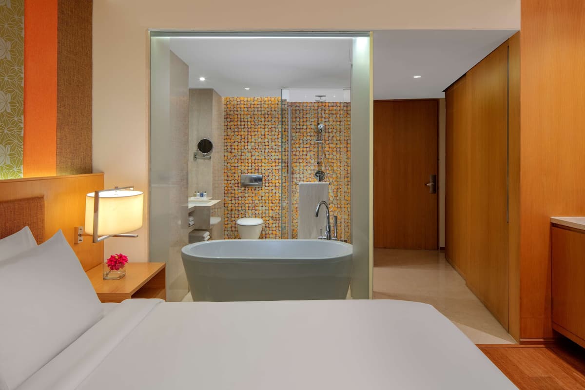 Ein modernes Hotelzimmer mit einem großen Bett, einer begehbaren Glasdusche, einem Holzschrank und einem kleinen Nachttischchen mit Lampe.