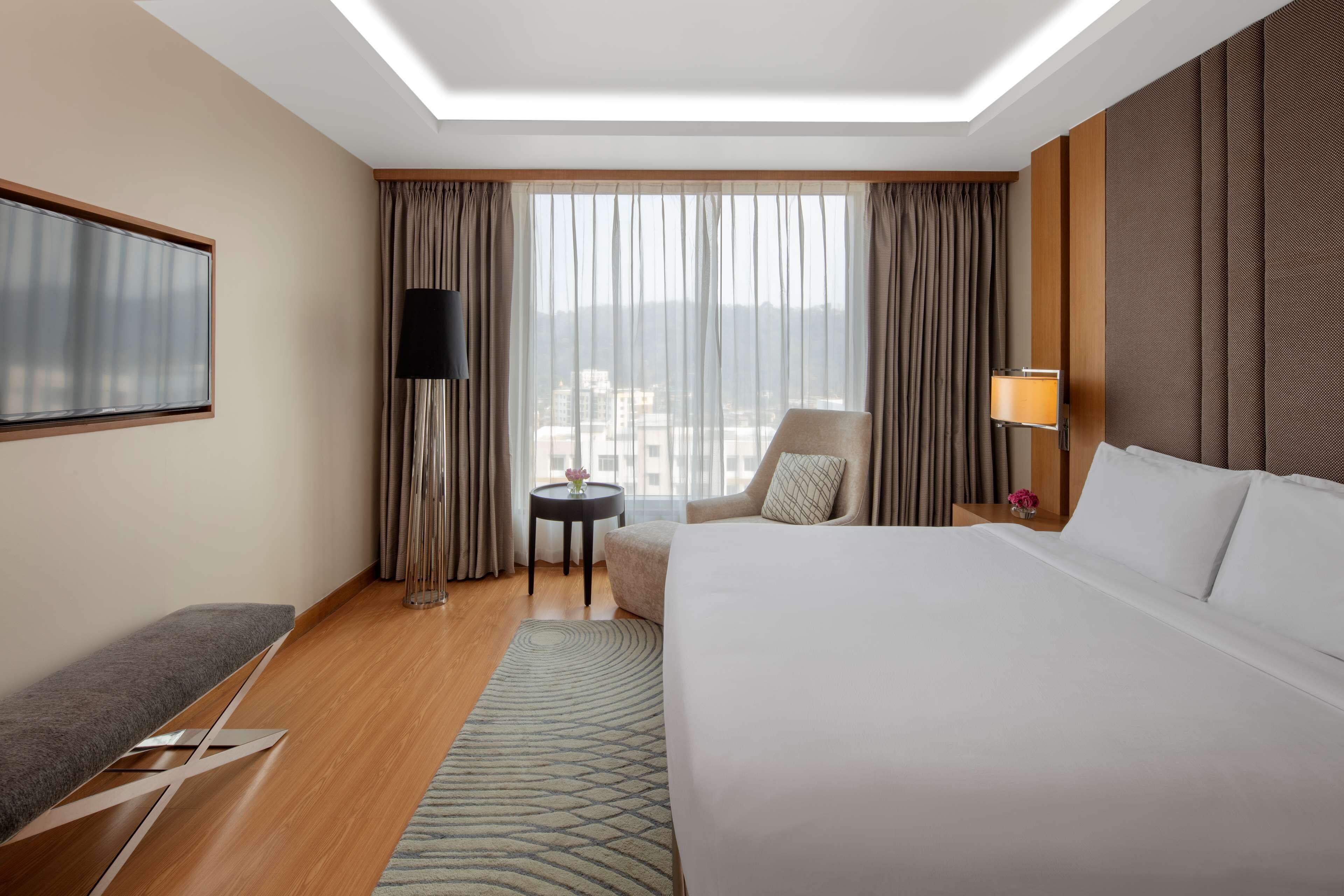 A hotel room with a large bed, a desk, a chair, a lamp, and a view of the city through the window.