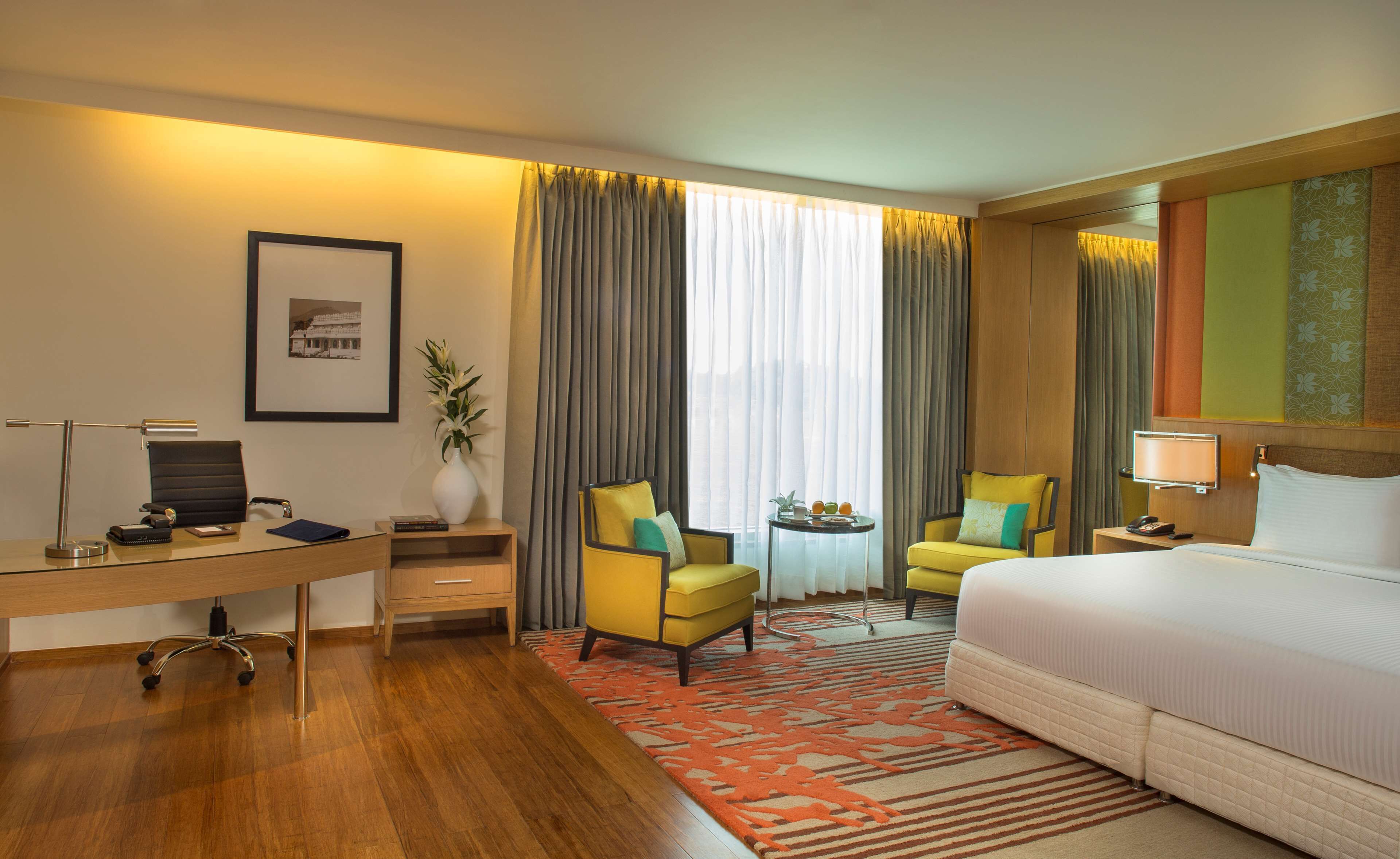 A hotel room with a bed, two chairs, a desk with a lamp, a small table with fruit, and a large window with curtains.