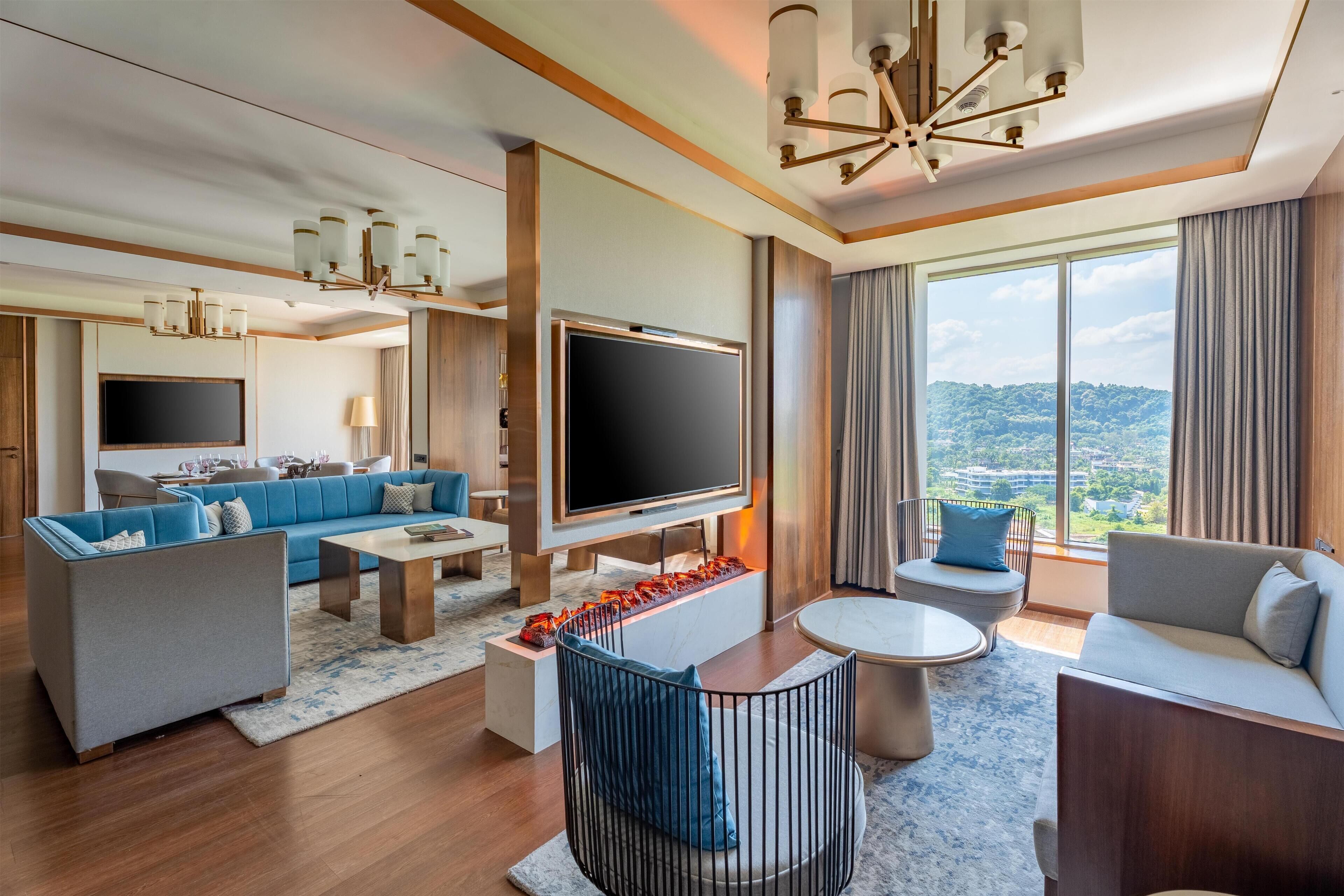 Une chambre d’hôtel moderne dotée d’un grand téléviseur à écran plat, d’un canapé bleu, d’un canapé gris, d’une table basse ronde et d’un coin repas avec une table et des chaises.