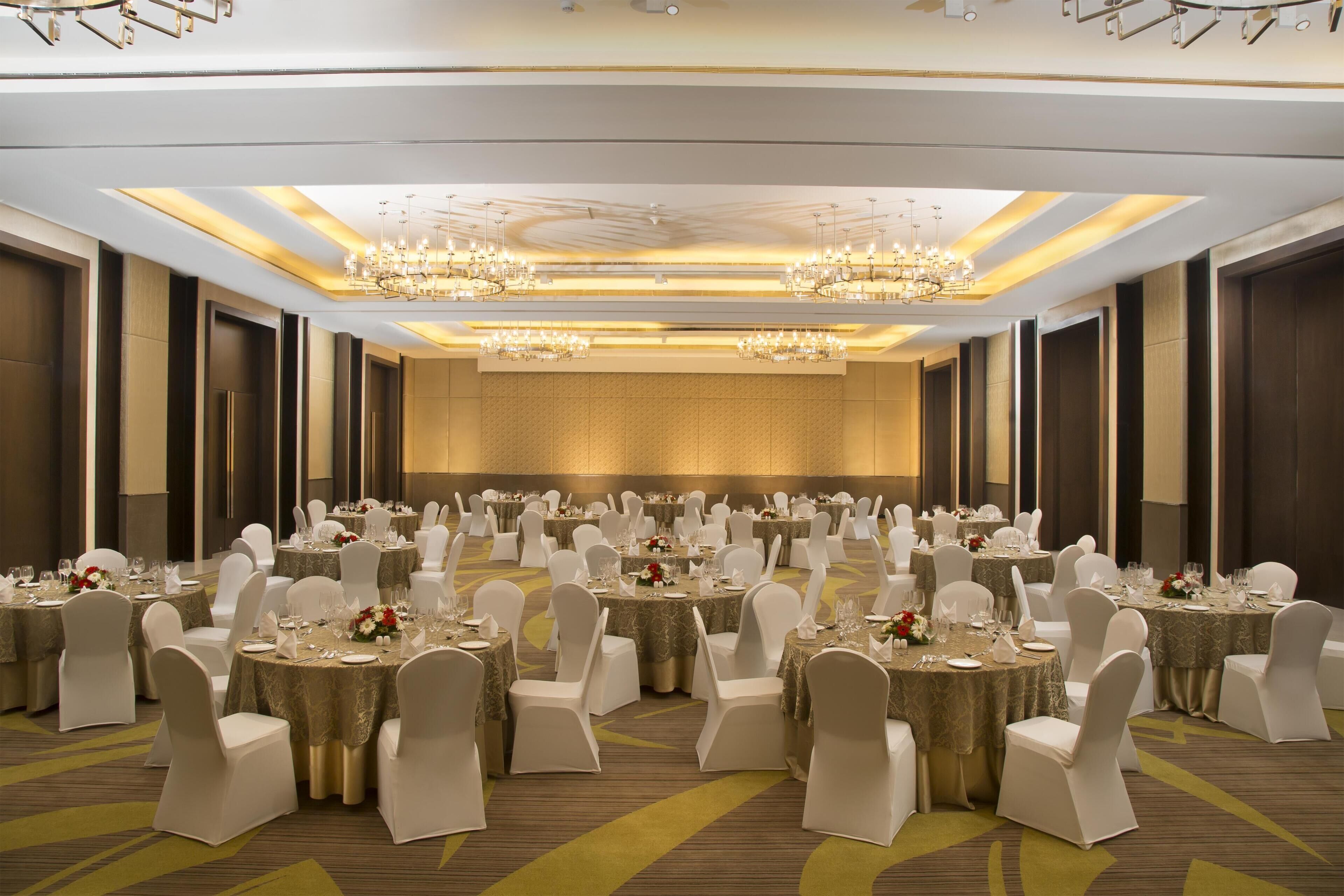 A large banquet hall with round tables, white chairs, chandeliers, and a carpeted floor.