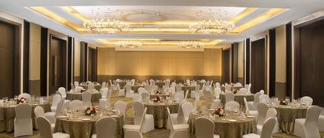 A large banquet hall with round tables, white chairs, chandeliers, and a carpeted floor.