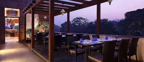 An outdoor dining area with wooden pergolas and tables, surrounded by chairs, and a view of trees and hills in the background.