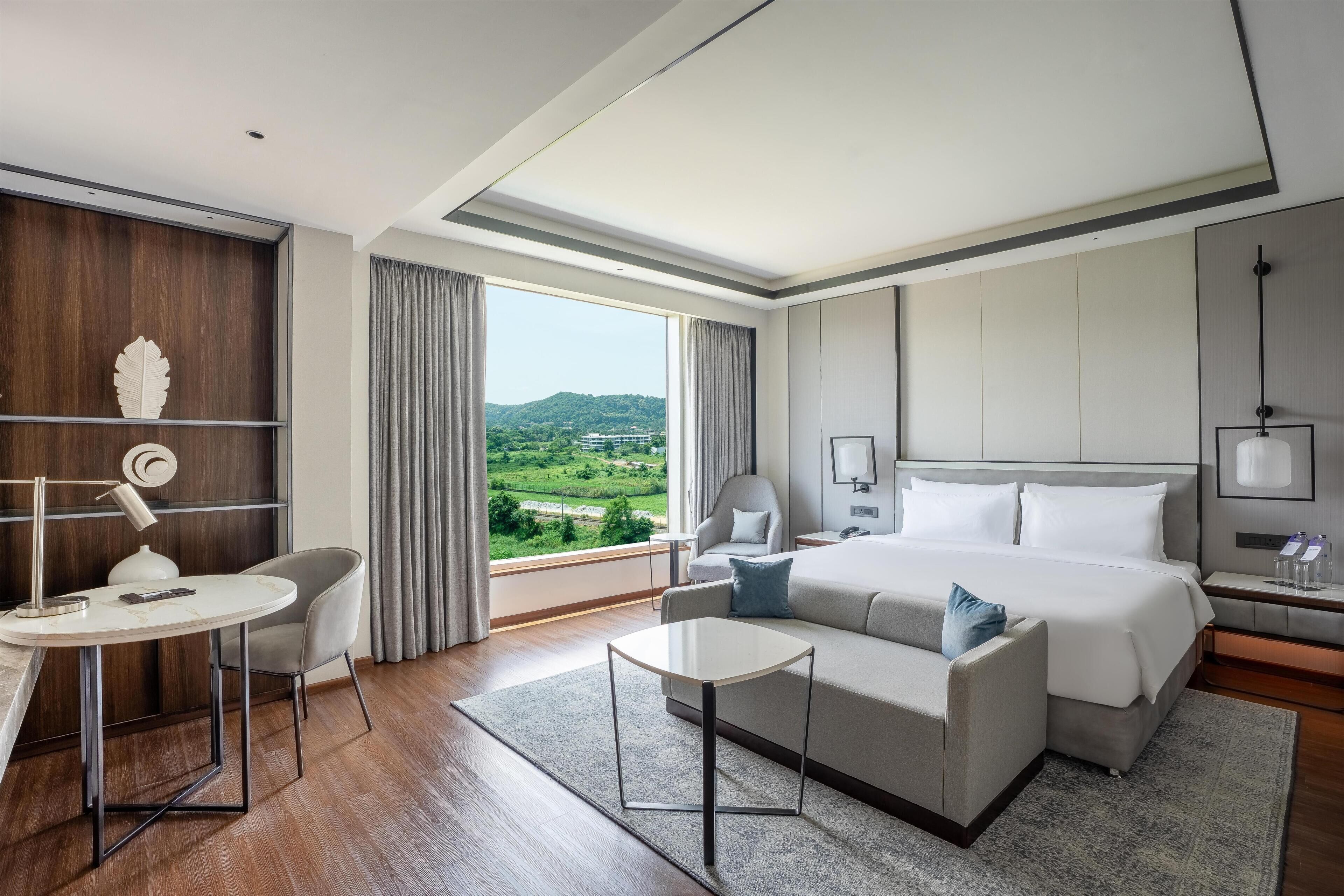 A hotel room with a large bed, a desk with a chair, a sofa, a view of the outdoors, and a modern design.