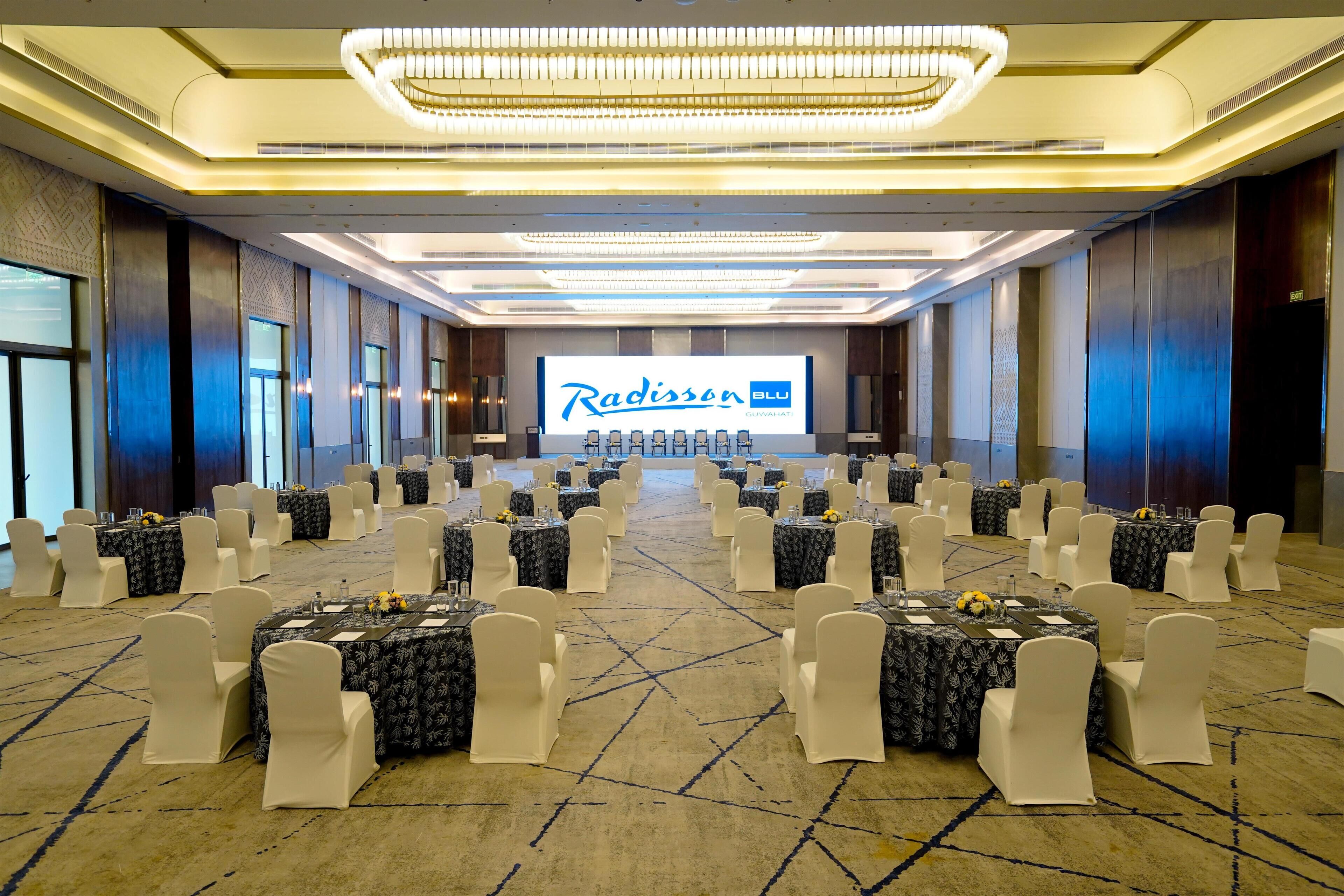 Une salle de conférence équipée de tables et de chaises, d’un grand écran affichant le logo « Radisson Blu », et d’un lustre.