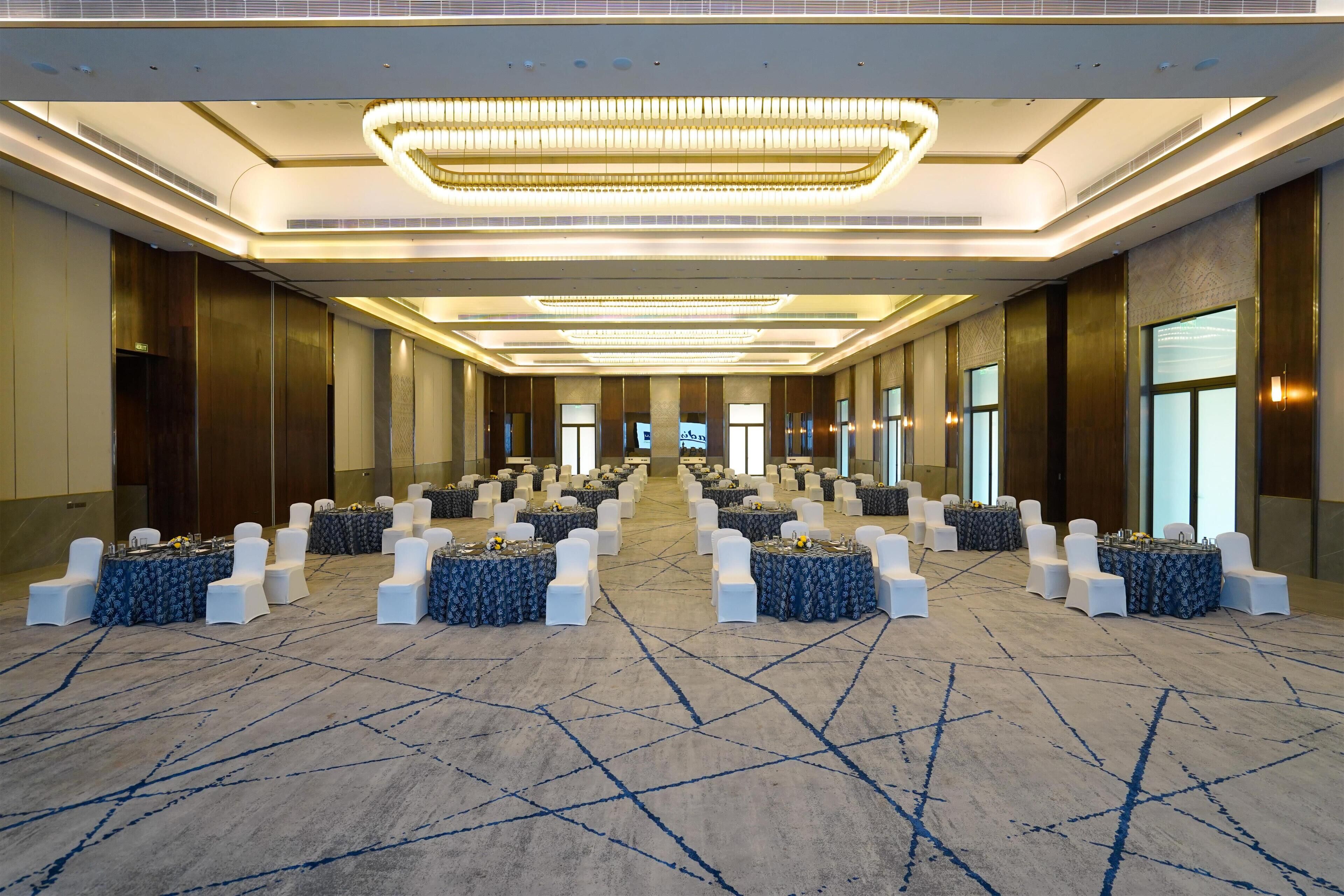 Une salle de conférence spacieuse, dotée de rangées de tables et de chaises, de grandes fenêtres et d’un lustre moderne.