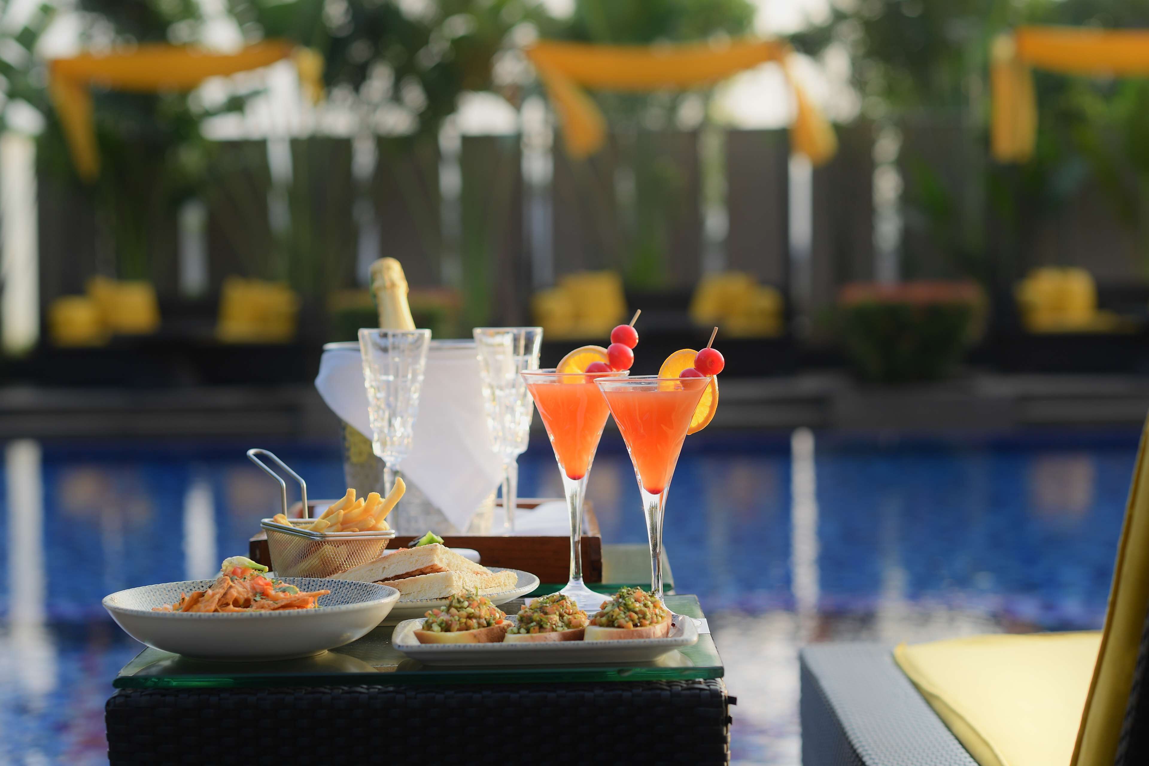 Una mesa con comida y bebidas junto a una piscina, con vistas despejadas al agua y la vegetación de fondo.