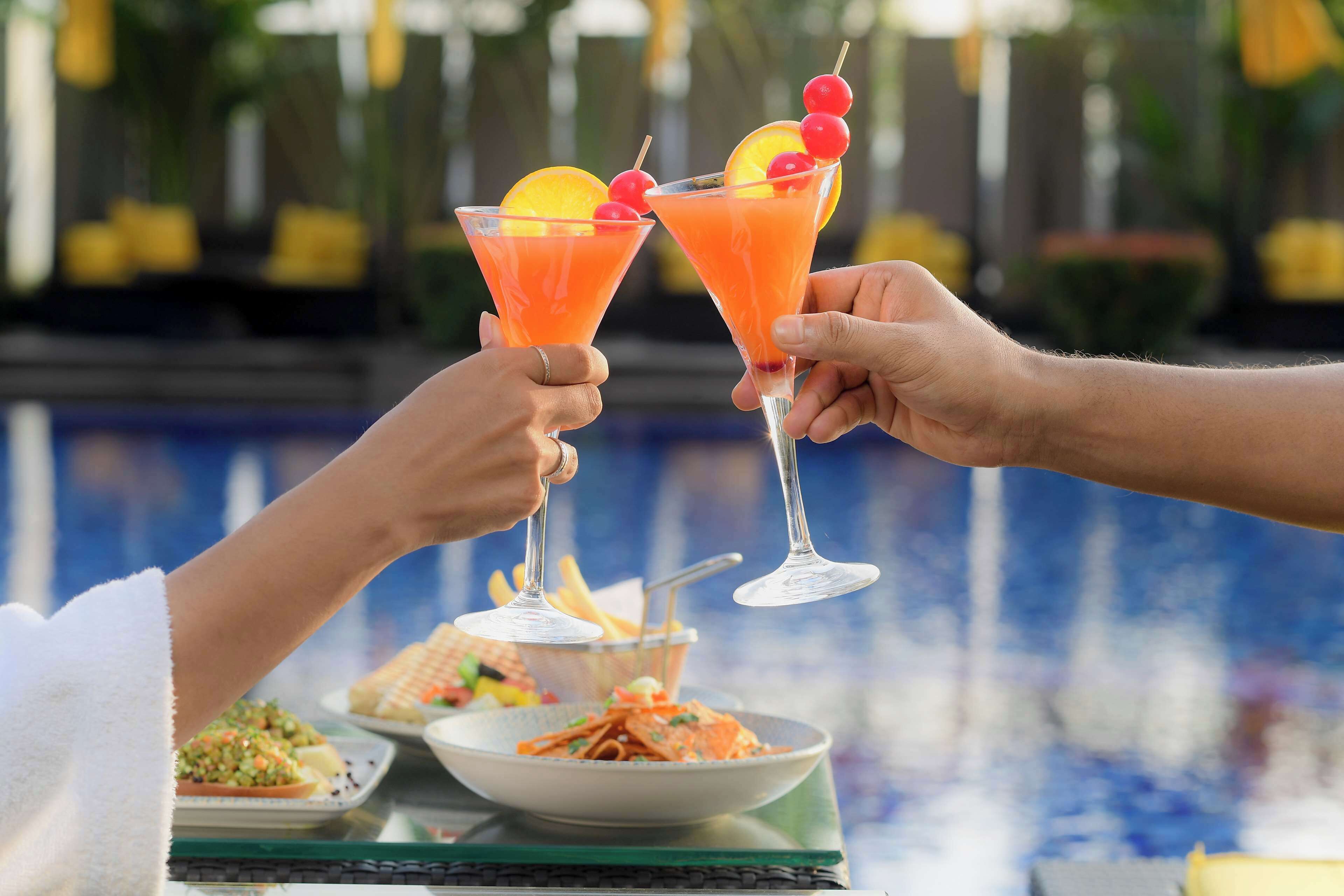Zwei Personen stoßen mit Cocktails an, die mit Früchten garniert sind. Im Hintergrund ein Pool und ein Tisch mit Speisen und Getränken.