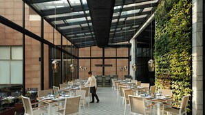Un restaurant au décor moderne, caractérisé par un plafond haut, une verrière et un mur végétal vertical. Les tables sont disposées avec soin et accompagnées de chaises, et on aperçoit un serveur qui se déplace.
