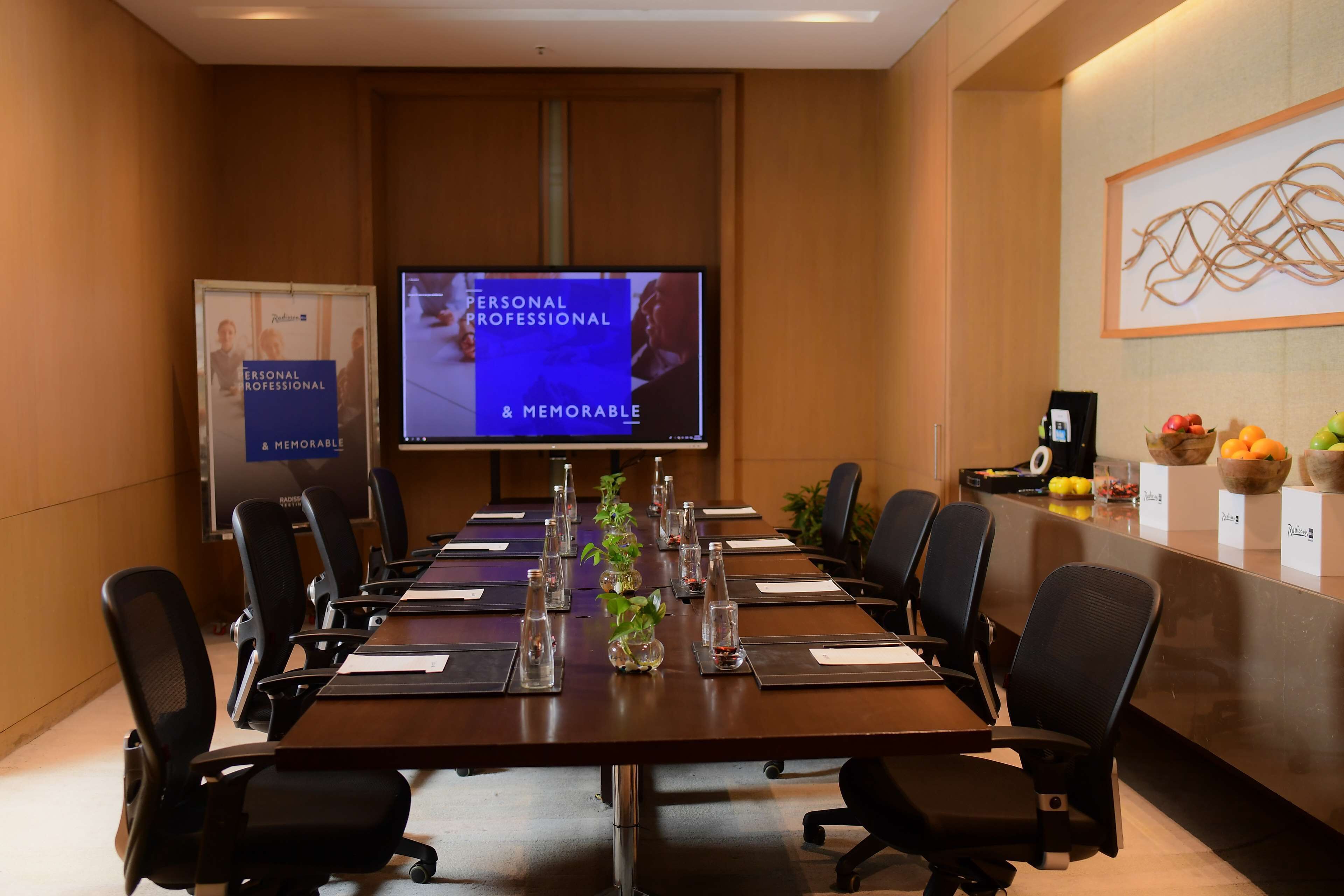 A conference room with a long table, black chairs, a TV screen, and a fruit basket on the counter.