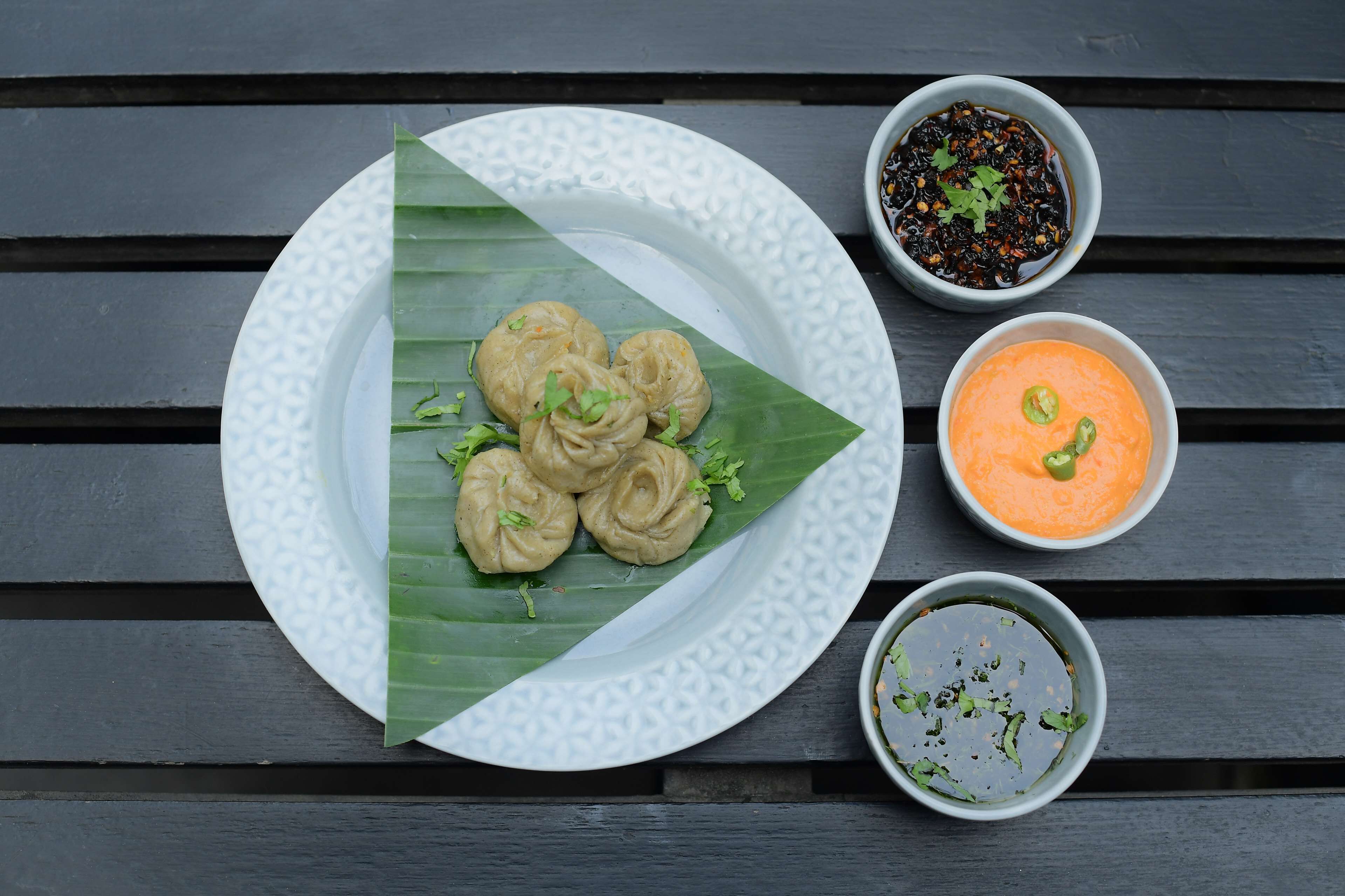 Un plato de dumplings sobre un plato blanco, acompañado de una hoja y tres recipientes pequeños con salsas para mojar.