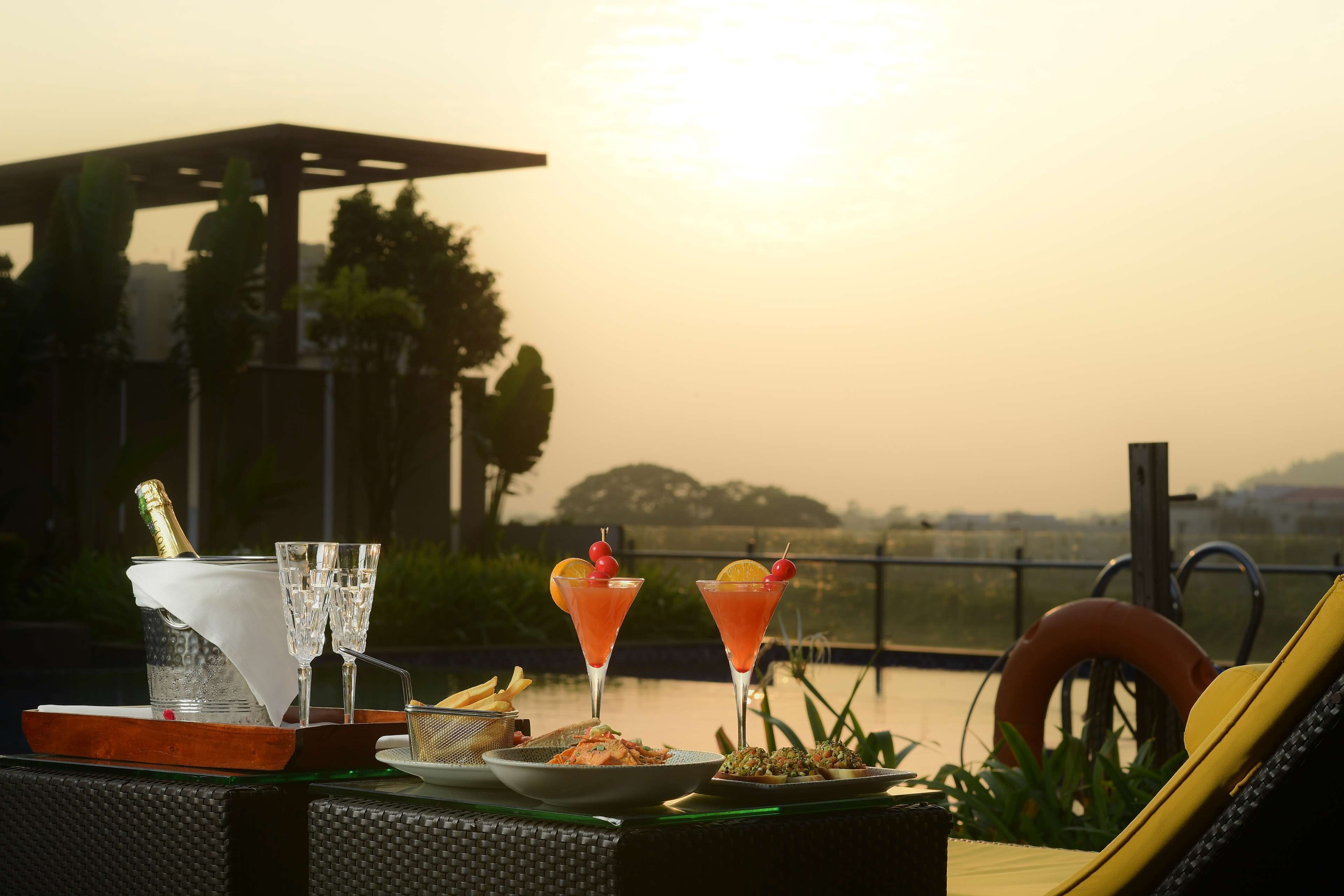 A table set with two cocktails, a bottle of champagne, and plates of food on a terrace overlooking a body of water.