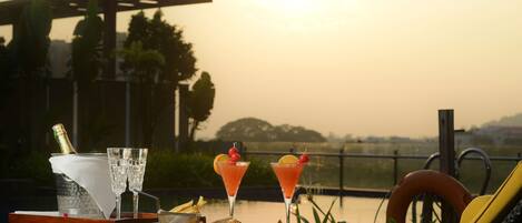 A table set with two cocktails, a bottle of champagne, and plates of food on a terrace overlooking a body of water.