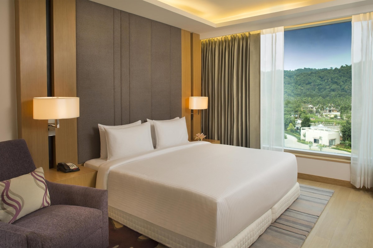 Ein Hotelzimmer mit einem großen Bett, zwei Nachttischlampen, einem Sessel, einem Nachttisch und einem Fenster mit Blick auf eine grüne Landschaft.