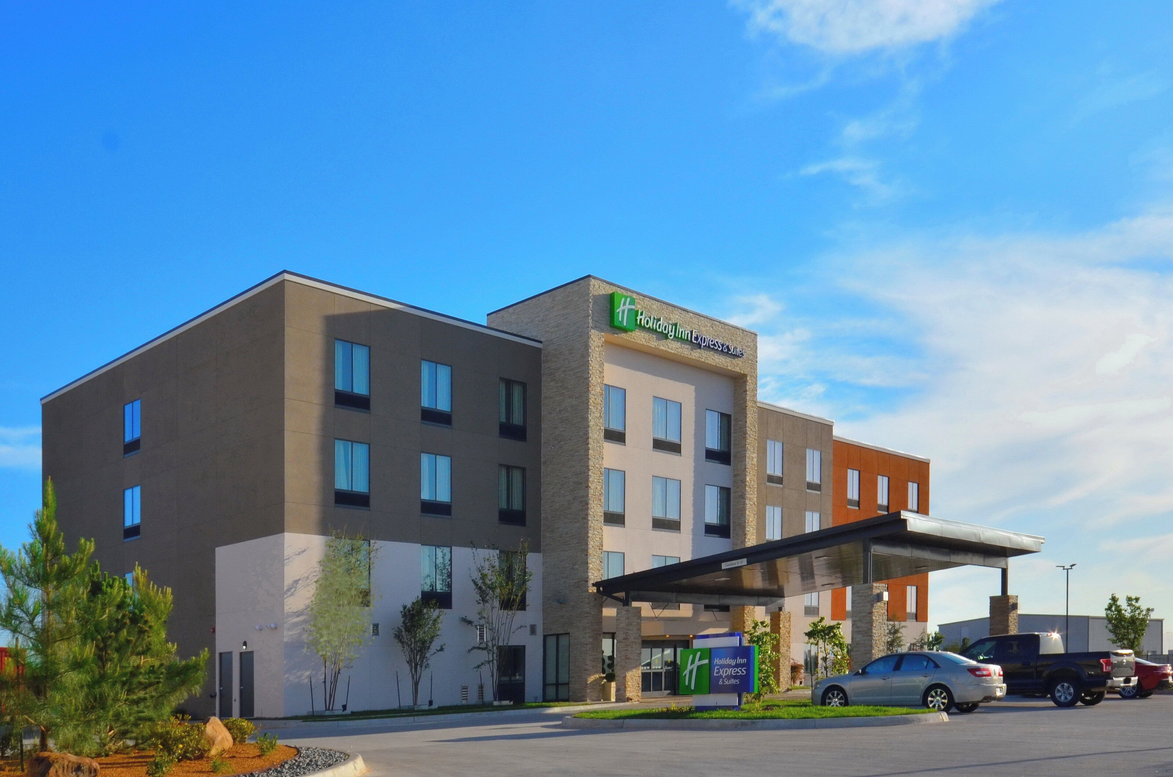 Un bâtiment de la chaîne Holiday Inn Express & Suites, doté d’un design moderne, comprenant un espace de stationnement couvert et offrant une vue sur un ciel dégagé.