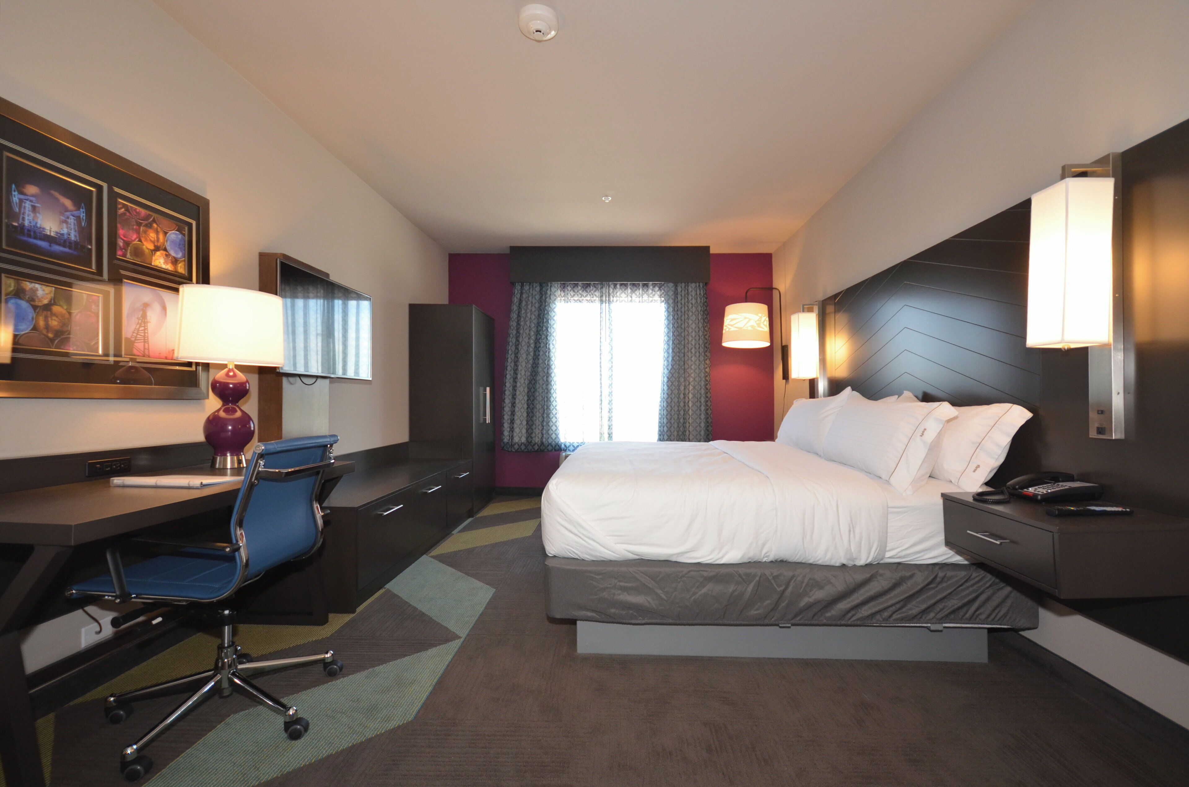 A hotel room with a bed, desk, chair, and a large window.