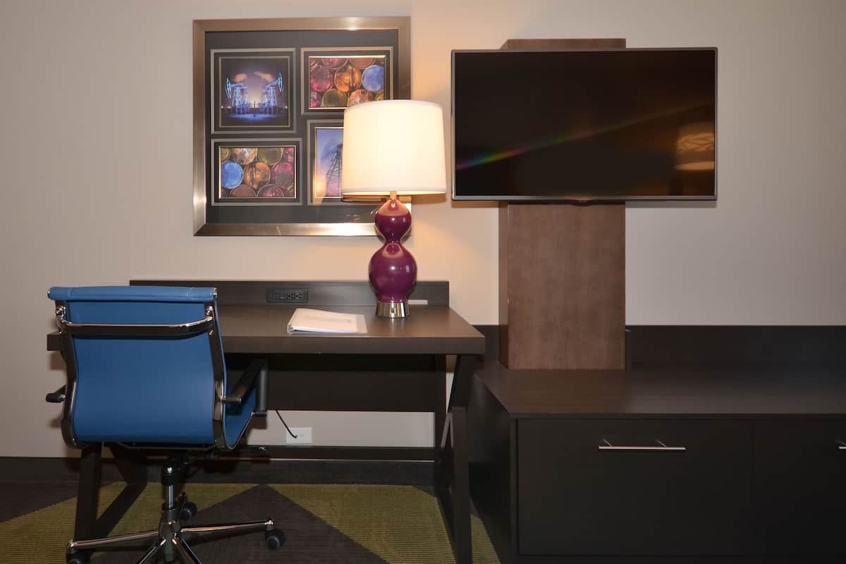 A hotel room with a desk, a blue office chair, a purple lamp, a flat-screen TV, and a wall-mounted artwork.