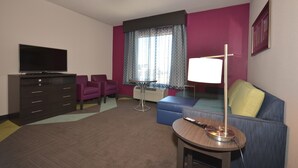 A hotel room with a flat-screen TV, a sofa, a small table with a lamp, a ceiling fan, and a window with patterned curtains. - Holiday Inn Express & Suites Oklahoma City Mid - Arpt Area by IHG (Oklahoma City)