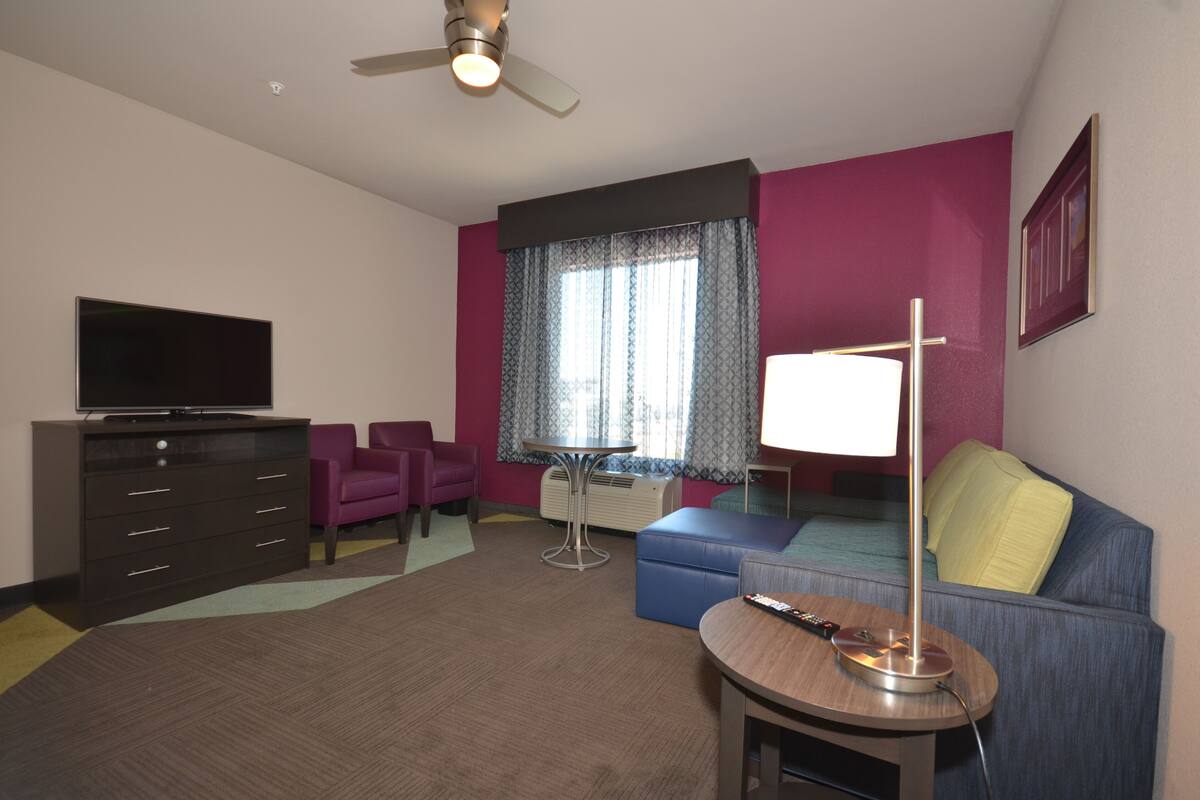 A hotel room with a flat-screen TV, a sofa, a small table with a lamp, a ceiling fan, and a window with patterned curtains.
